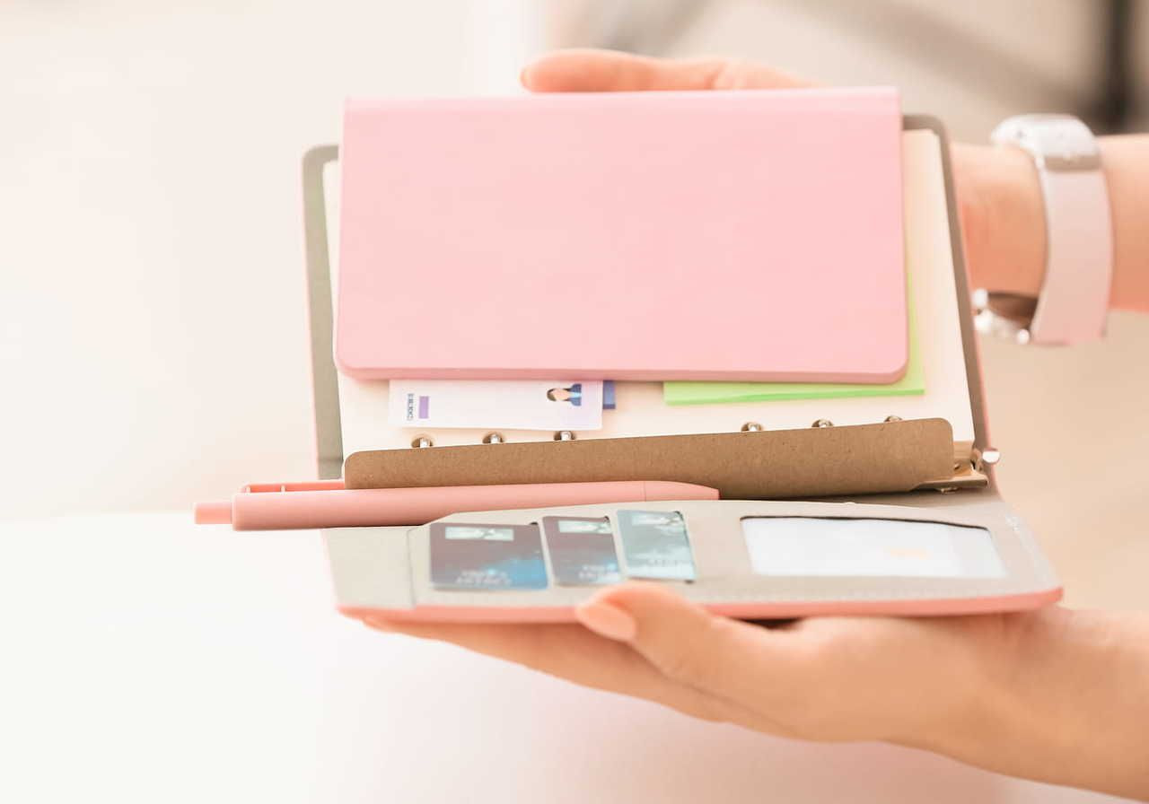 Hands holding an open pink planner containing SD cards, a closed notebook, and documents. A pink pen rests on the planner.