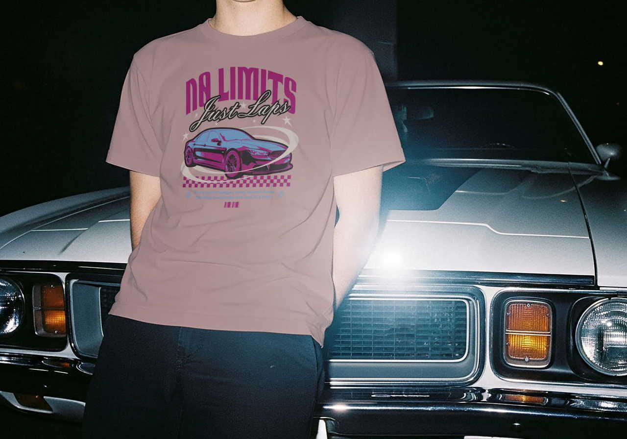 A man wearing a pink "No Limits Just Laps" t-shirt, featuring a graphic of a racing car. He stands next to a vintage car at night.