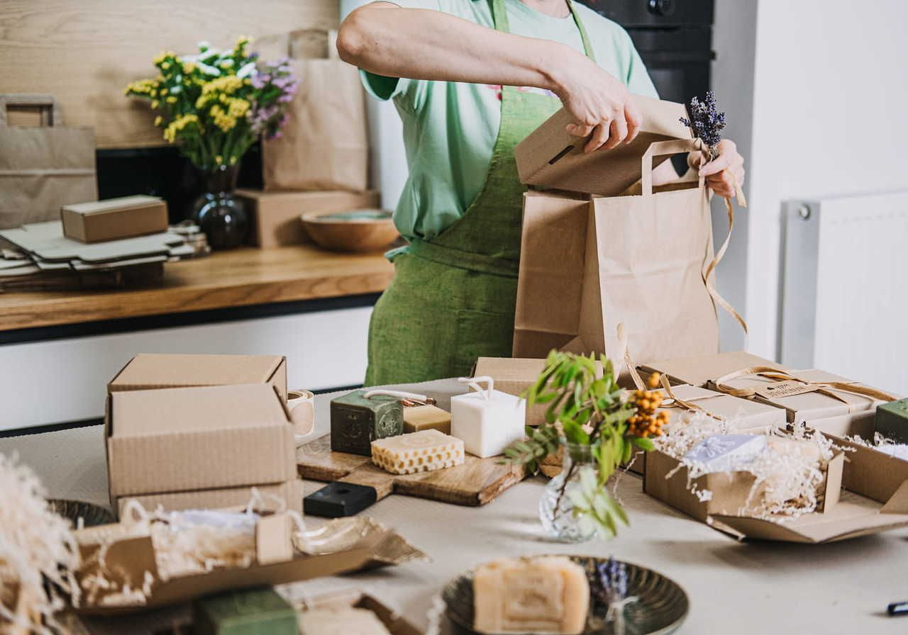 Person in a green apron packing handmade soaps and candles into a brown paper bag on a wooden table, surrounded by flowers and gift boxes.