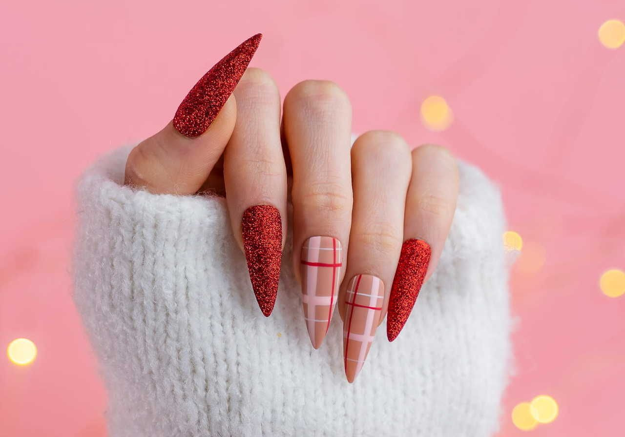 A hand with long, pointed nails featuring a combination of glittery red and a nude design with pink stripes, against a light pink background.