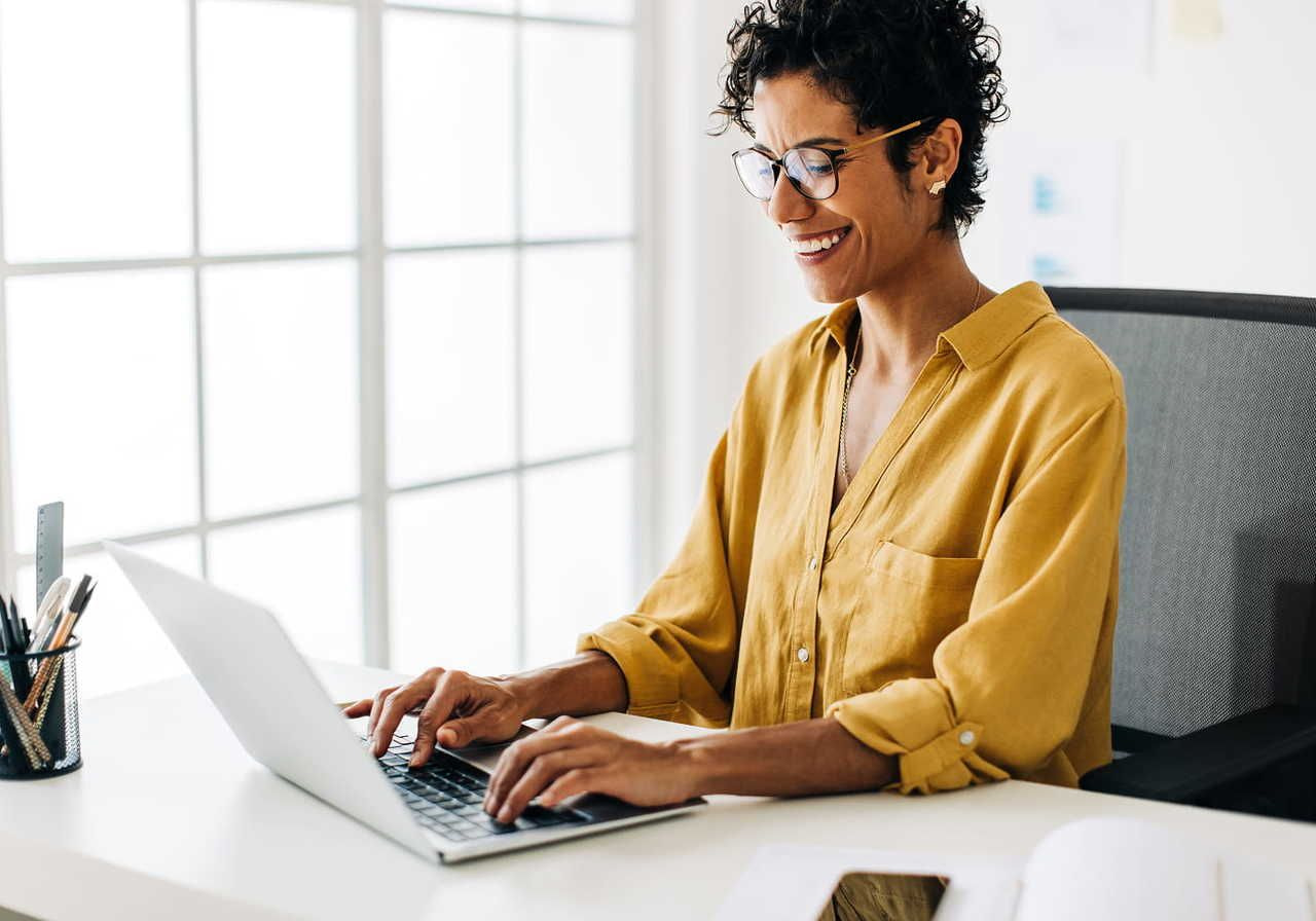 A smiling woman in glasses, wearing a yellow sweater, is using a laptop in a home office to compare Gelato vs Printify.