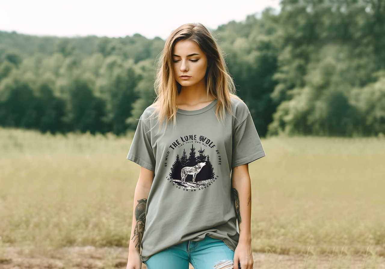 Young woman with long hair stands in a grassy field, wearing a gray t-shirt featuring a wolf and trees graphic.