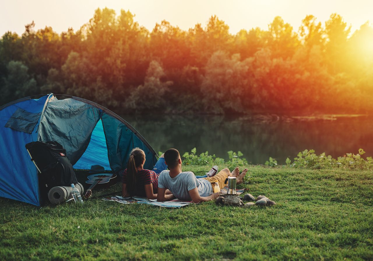 A couple relaxes on a blanket near a blue tent by a serene lake. 