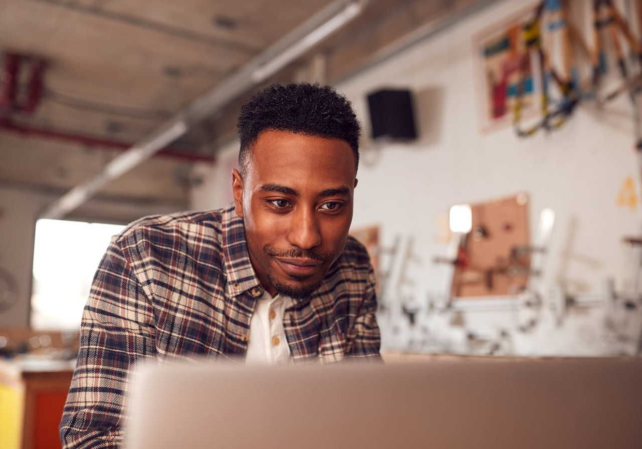 A man in a flannel shirt is focused on a laptop screen, researching dropshipping business ideas.