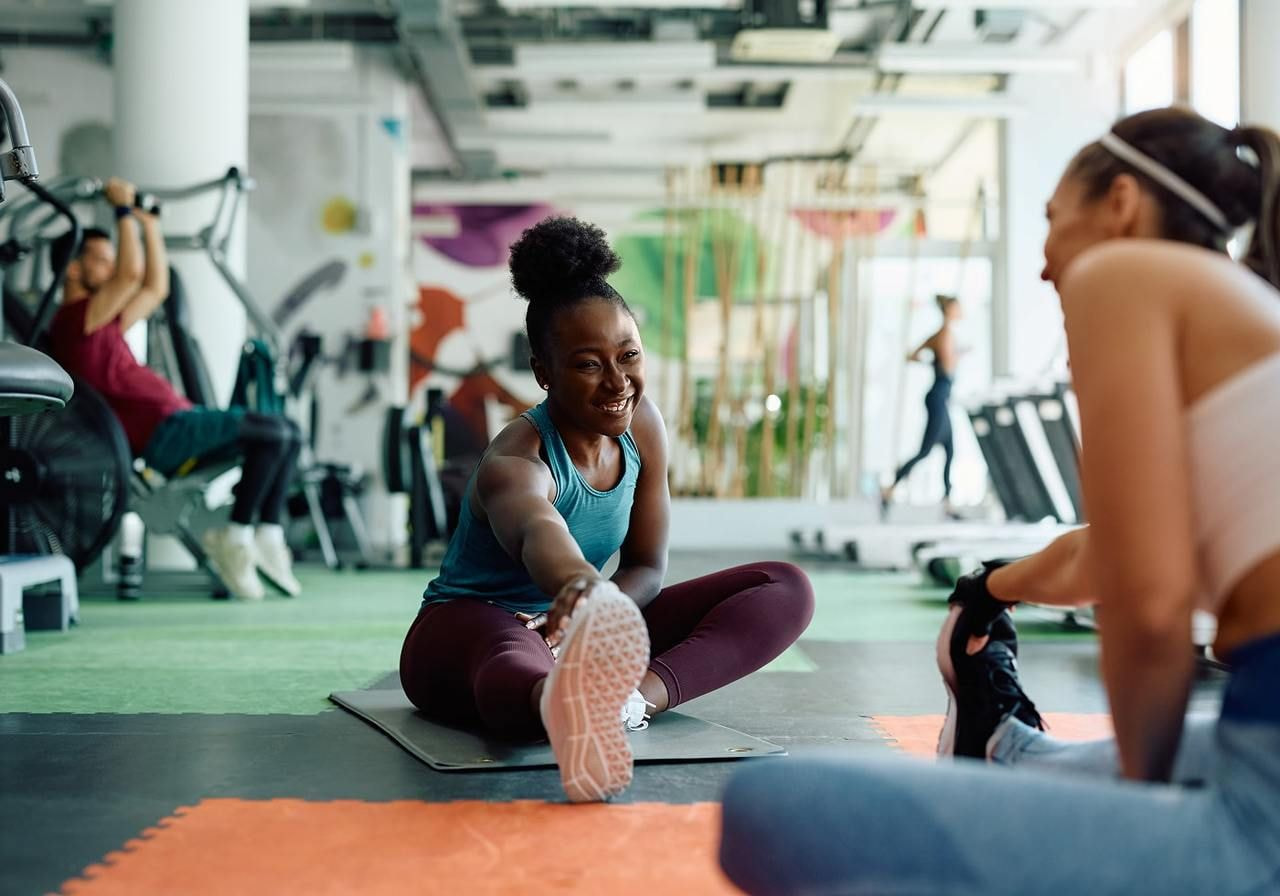 A smiling woman is stretching in a gym while talking to another woman.