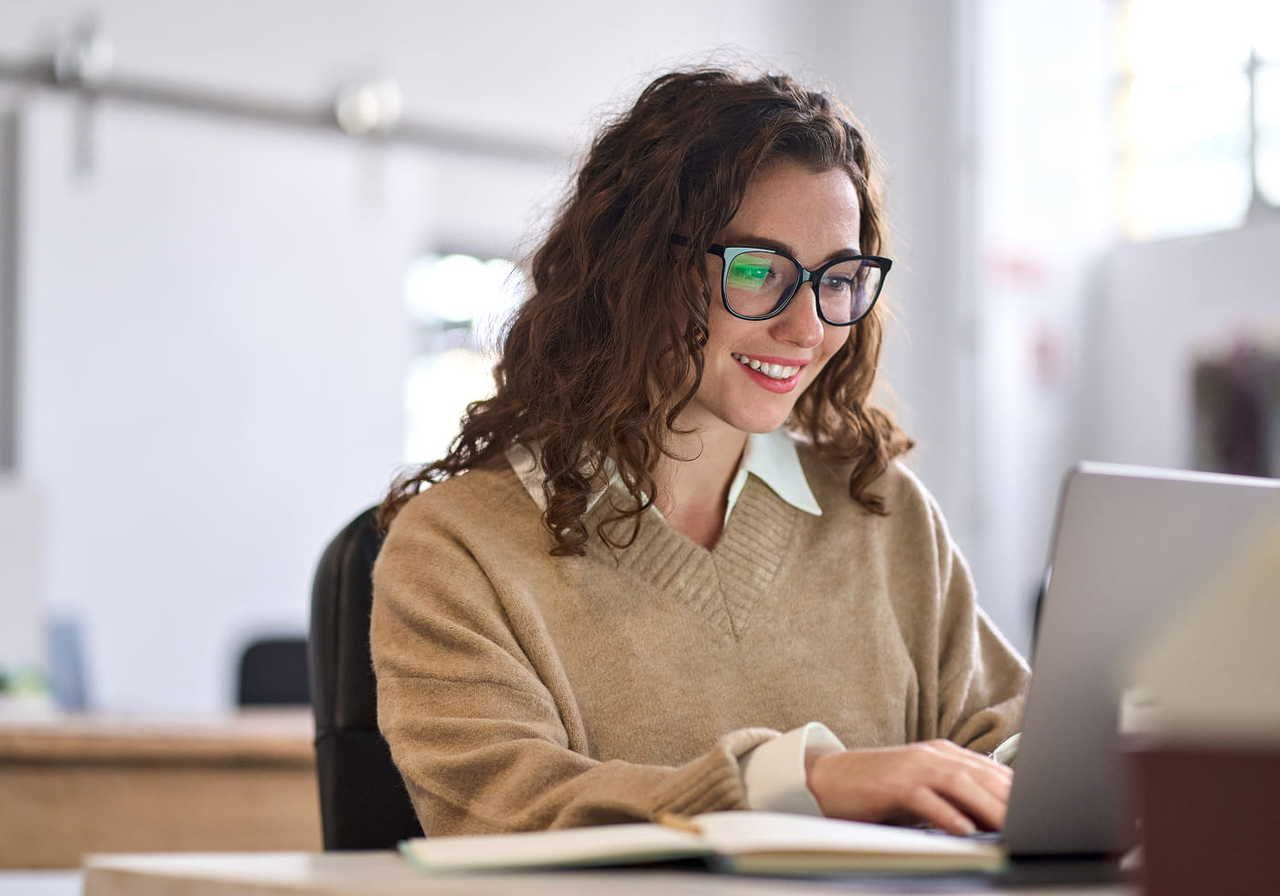 A woman with curly hair and glasses is sitting at a desk, smiling while typing on a laptop. She is wearing a beige sweater.