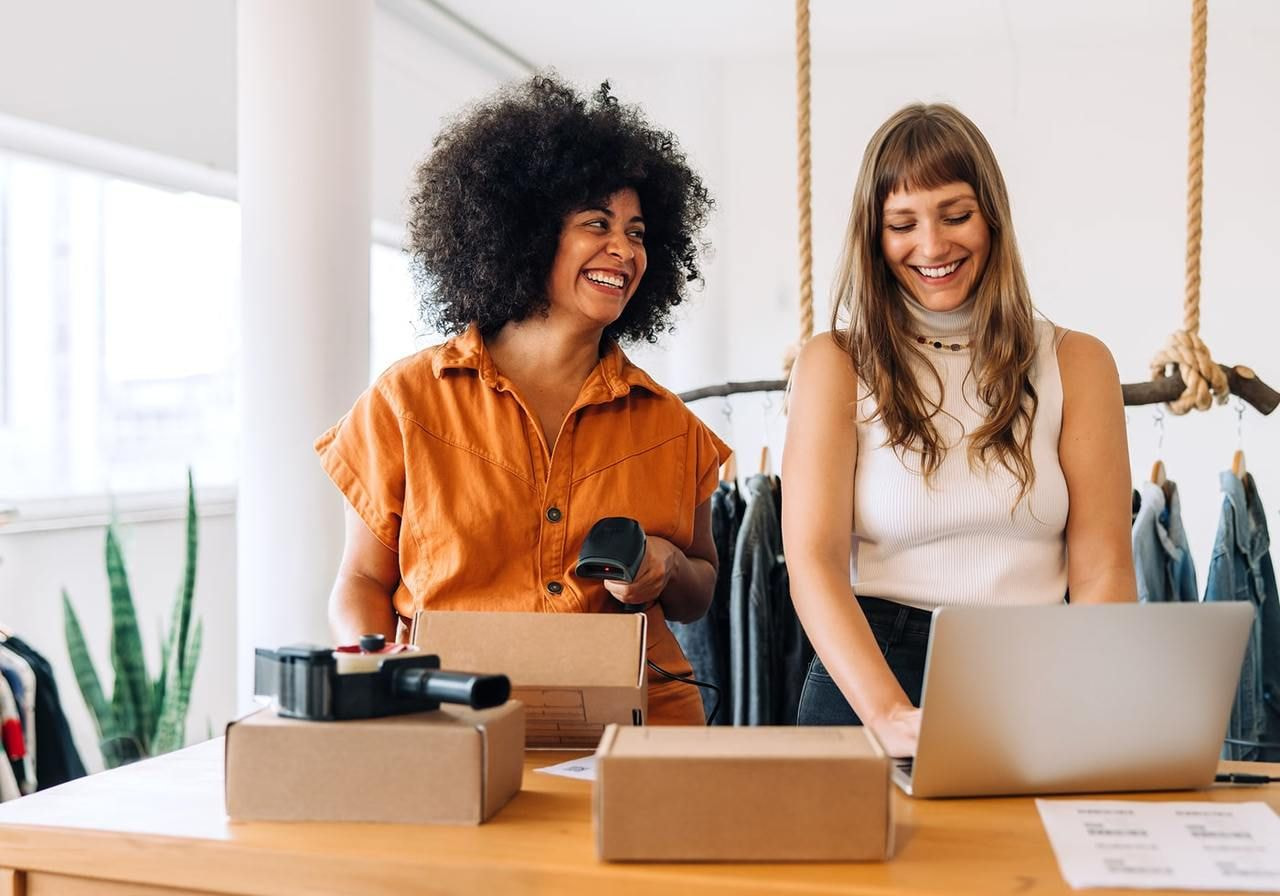 Two women collaborate in an office surrounded by boxes and a laptop, engaged in a discussion.