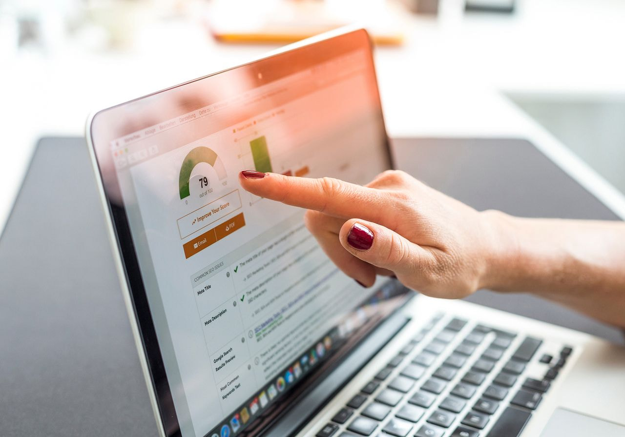 A person with red nail polish points at a laptop screen displaying analytics, featuring a green dial and bar graphs, in a bright office setting.