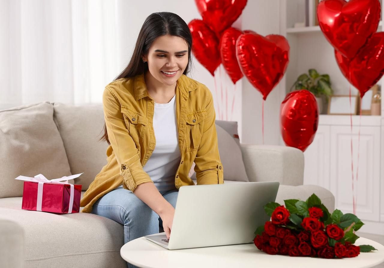 A smiling woman is using a laptop, searching for Valentine’s marketing ideas, on a couch with heart-shaped balloons, a gift, and roses nearby.