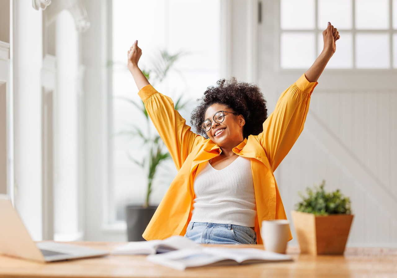 A woman with curly hair and glasses, wearing an orange shirt, sits at a table, her arms raised in a joyful pose. Books and a laptop in front of her.