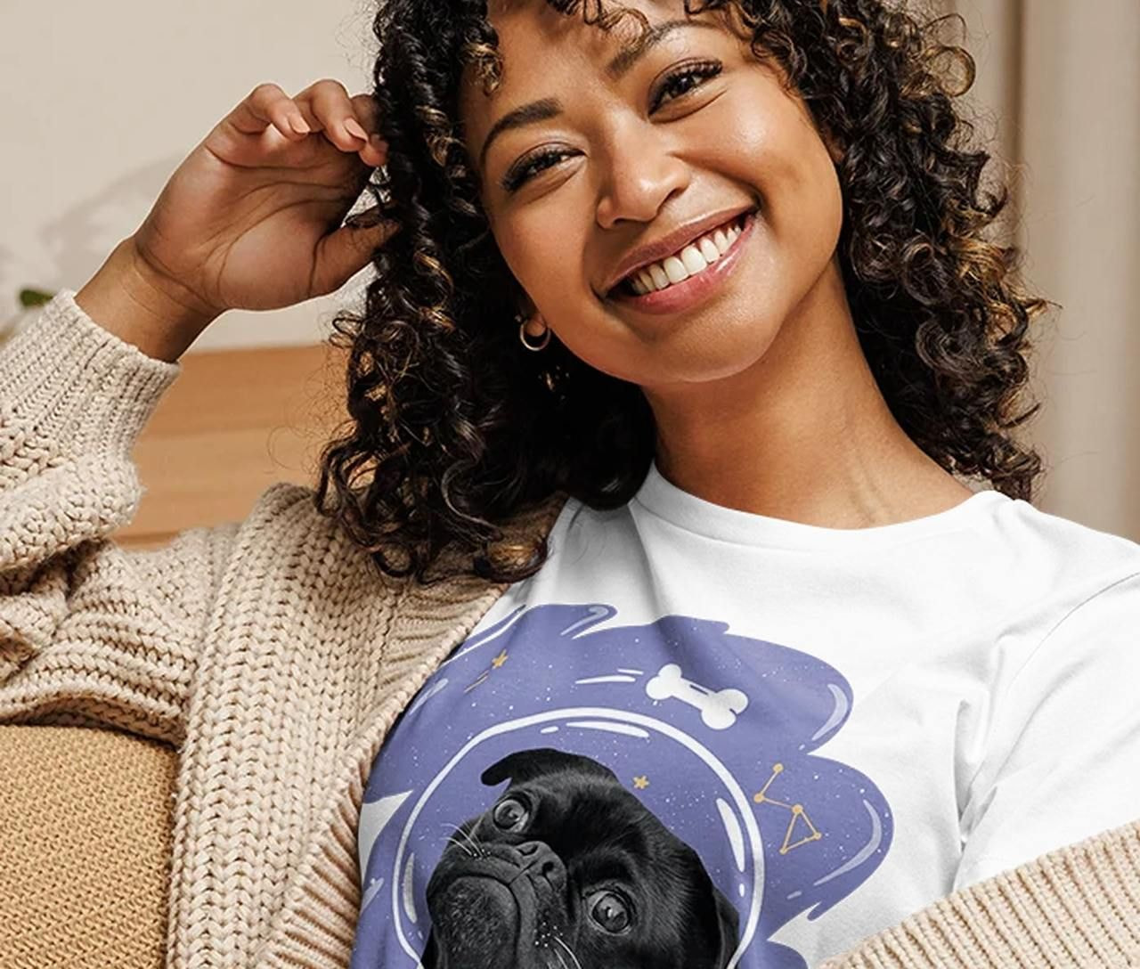 A woman with curly hair is wearing a cozy beige cardigan and a white t-shirt featuring a pug in a space-themed design.