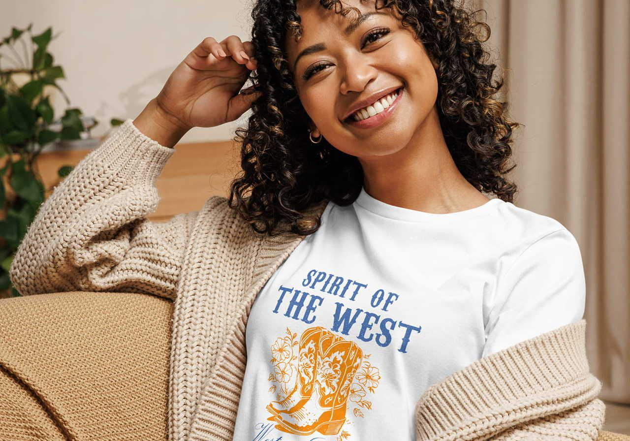 Smiling woman seated on a beige couch, wearing a cozy knitted cardigan and a white t-shirt with "Spirit of the West" and a cowboy boot design.