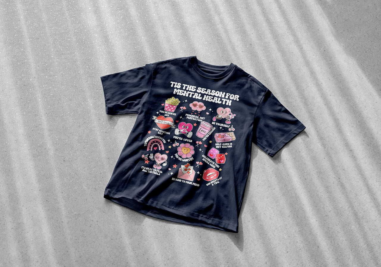 A black t-shirt on a textured gray surface features colorful, positive cartoon graphics and text reading, "Tis the Season for Mental Health".