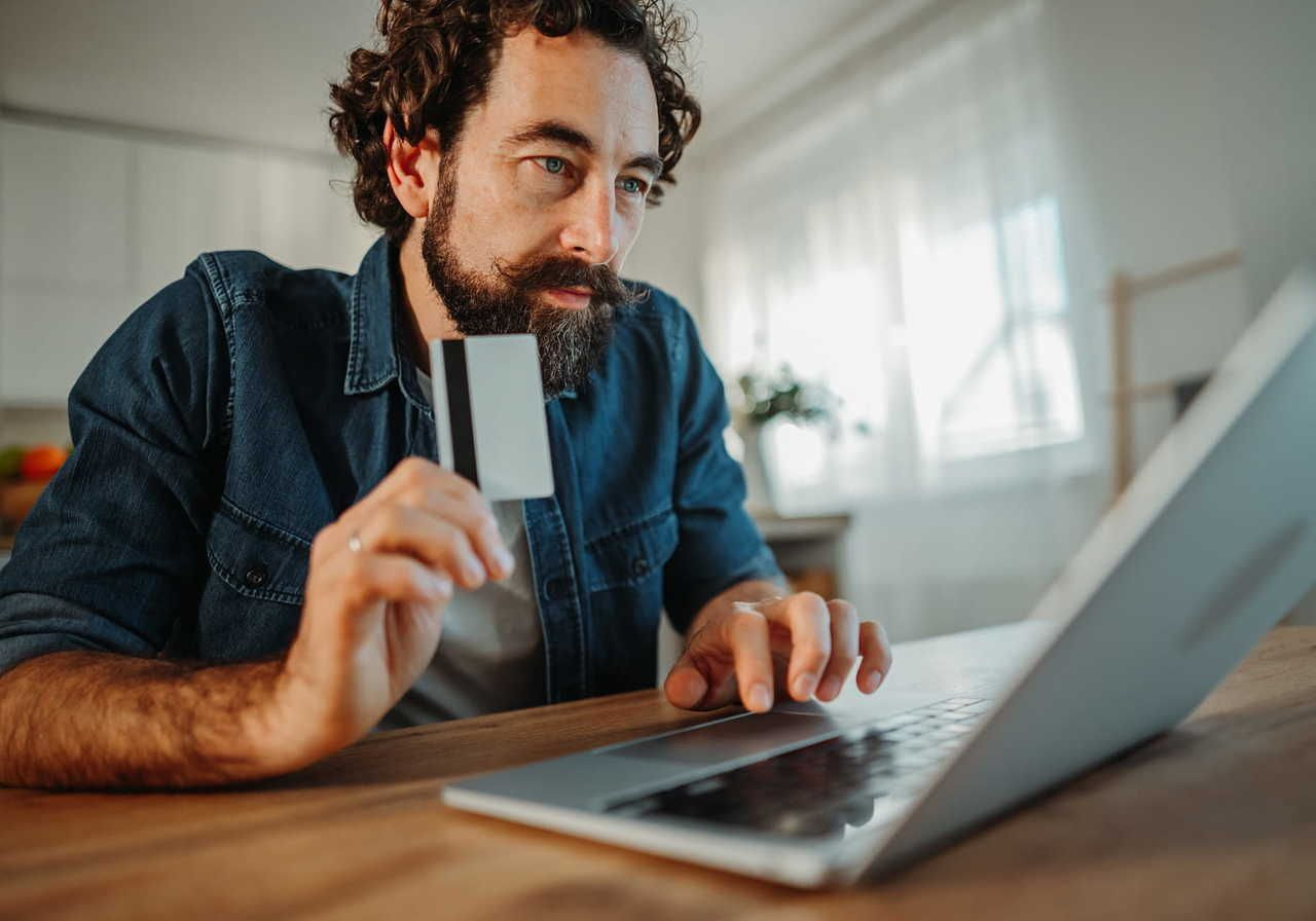 A man with curly hair and a beard is holding a credit card while using a laptop at a desk.