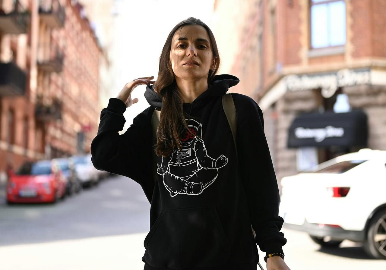 A woman in a black hoodie with an astronaut design stands confidently on a city street, with red brick buildings and parked cars in the background.