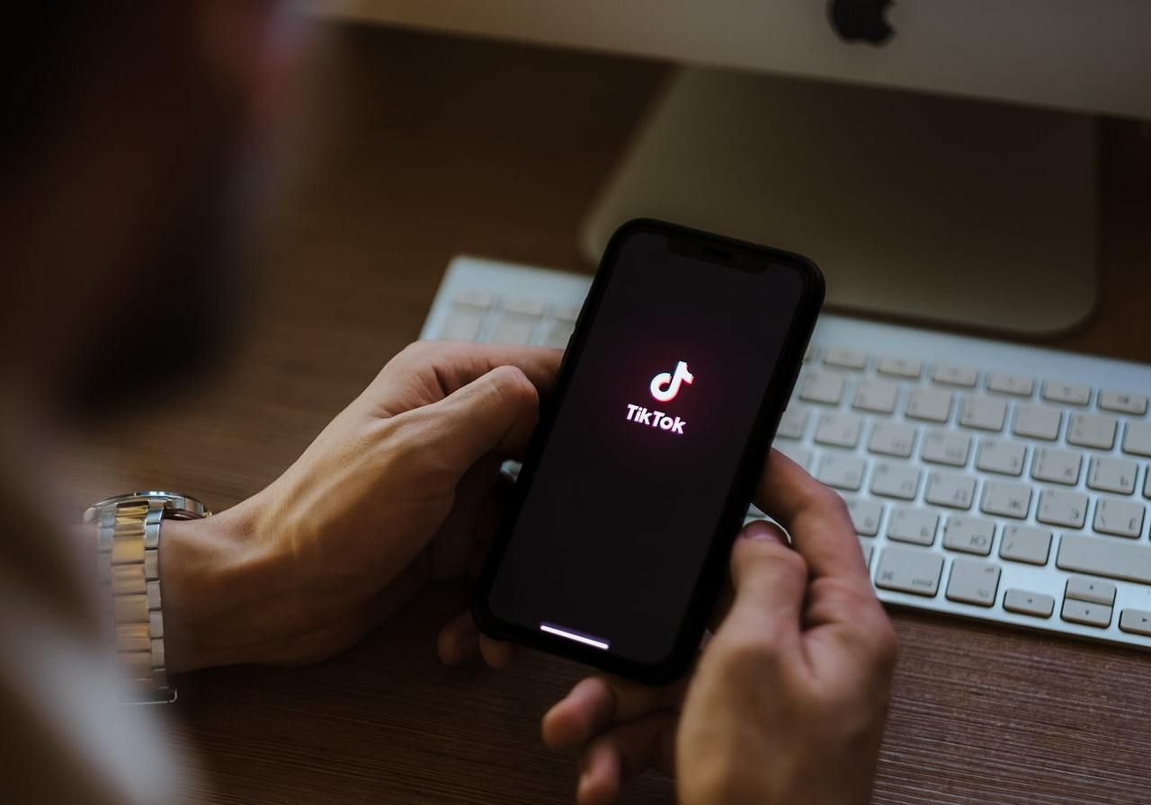 Hands holding a smartphone displaying the TikTok logo on the screen. Behind the desk, a white keyboard and monitor are visible.
