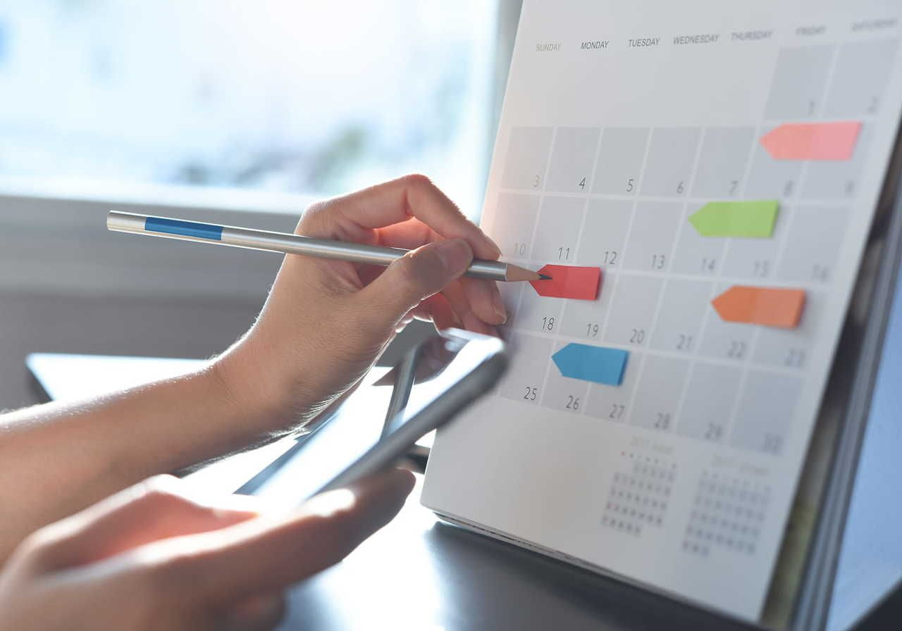 A person’s hands are pointing to a certain calendar date with a pen while holding a mobile phone in the other hand.