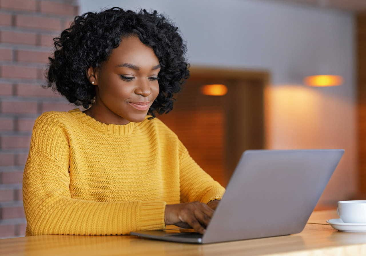 A woman wearing a yellow sweater smiles as she types on a laptop, with a coffee cup nearby.