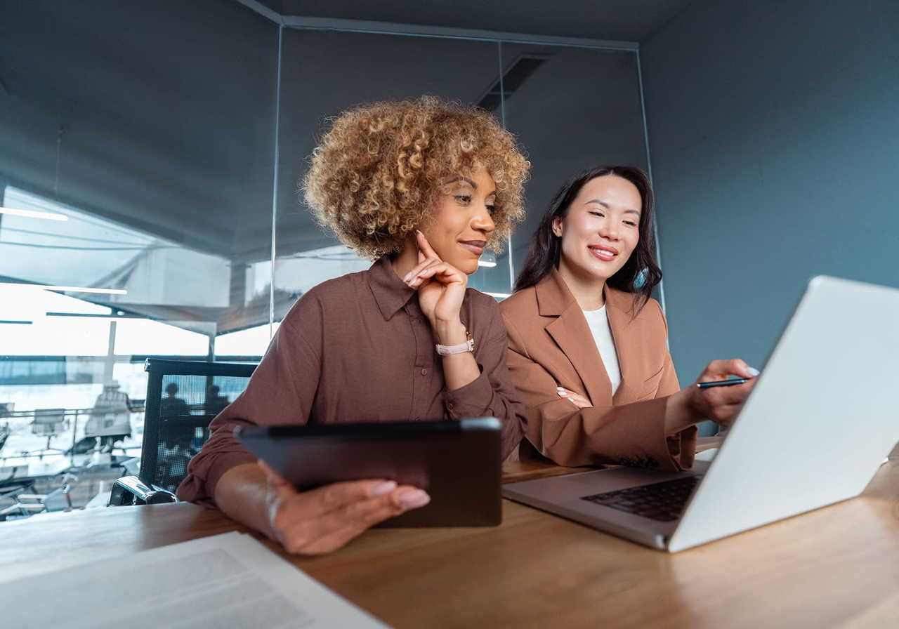 Two women working together with a laptop and a tablet in a modern office setting.