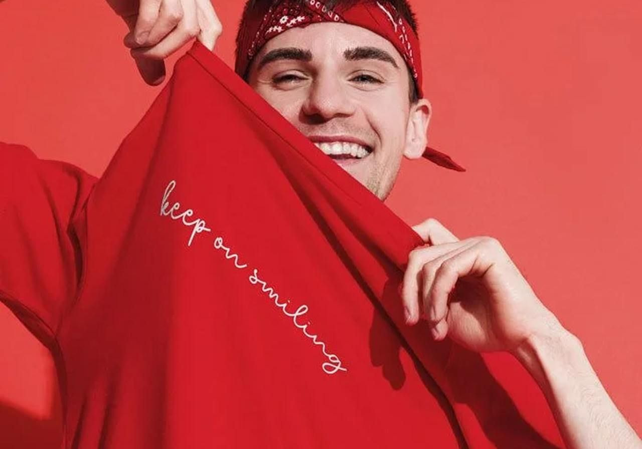 A smiling man in red, wearing a bandana, holds up a shirt with "keep on smiling" text. 