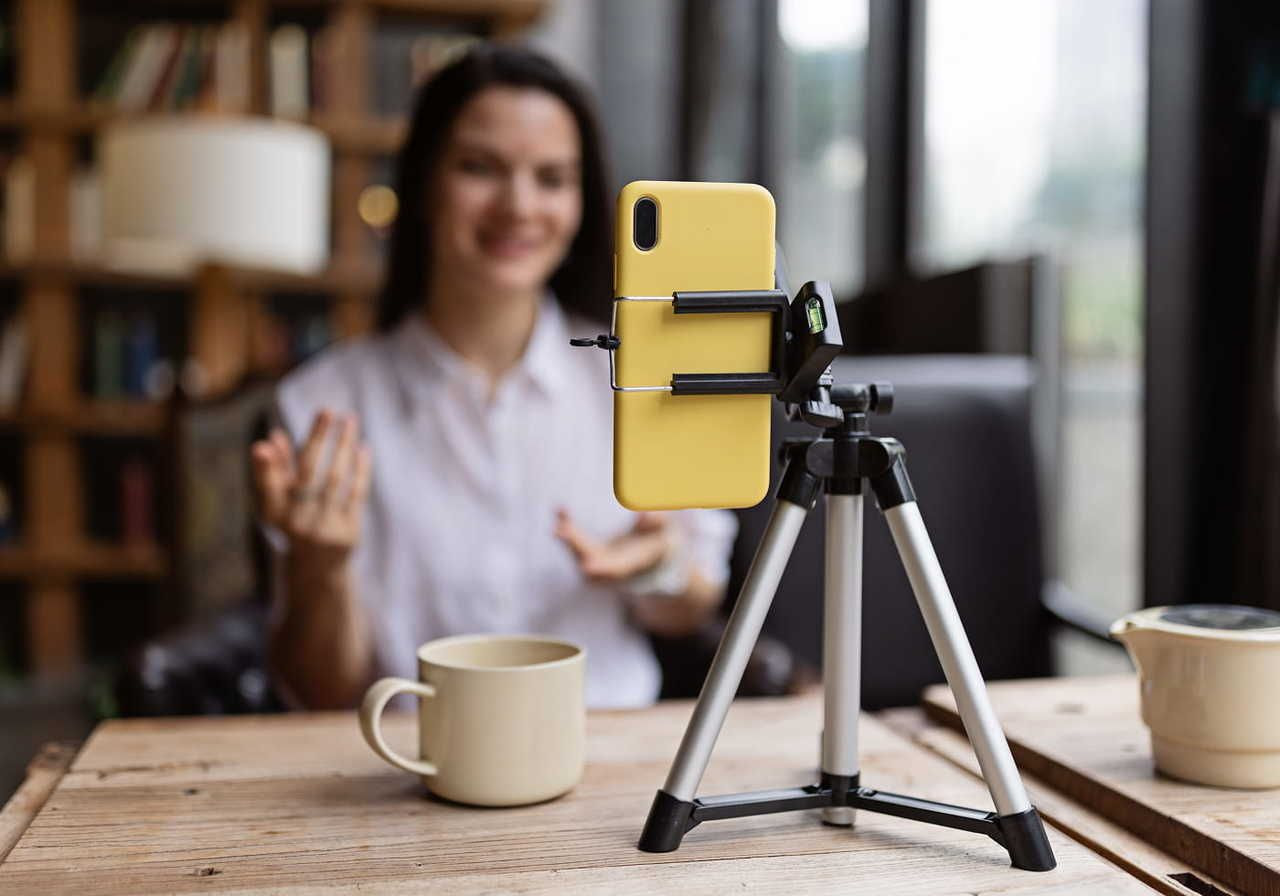 A woman is gesturing while recording herself with a smartphone on a tripod. A mug and a teapot are on the table.