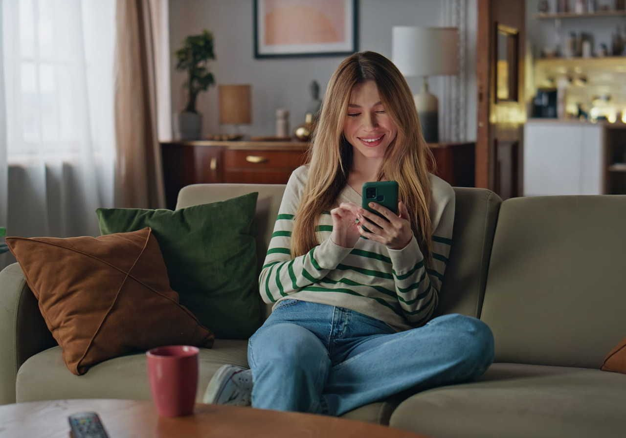 A young woman sitting on a sofa, smiling while using a smartphone, with a cup and decorative pillows nearby.