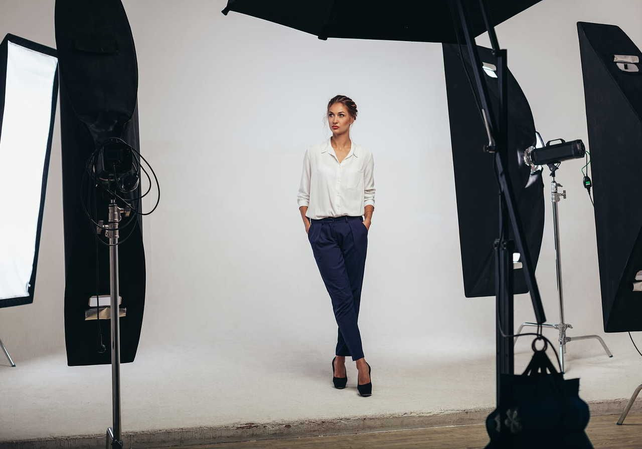 A model stands in a studio setting surrounded by lighting equipment, wearing a white blouse and dark pants.