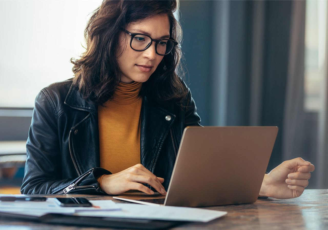 A woman wearing glasses is focused on her laptop, engaged in work at a desk.