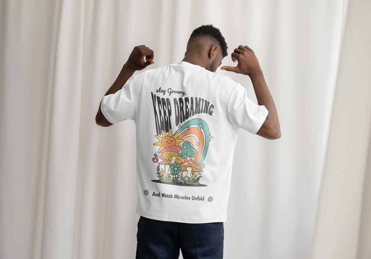 Man facing away, wearing a white t-shirt with a colorful retro design showing sun, mushrooms, rainbow, and the text "Keep Dreaming".