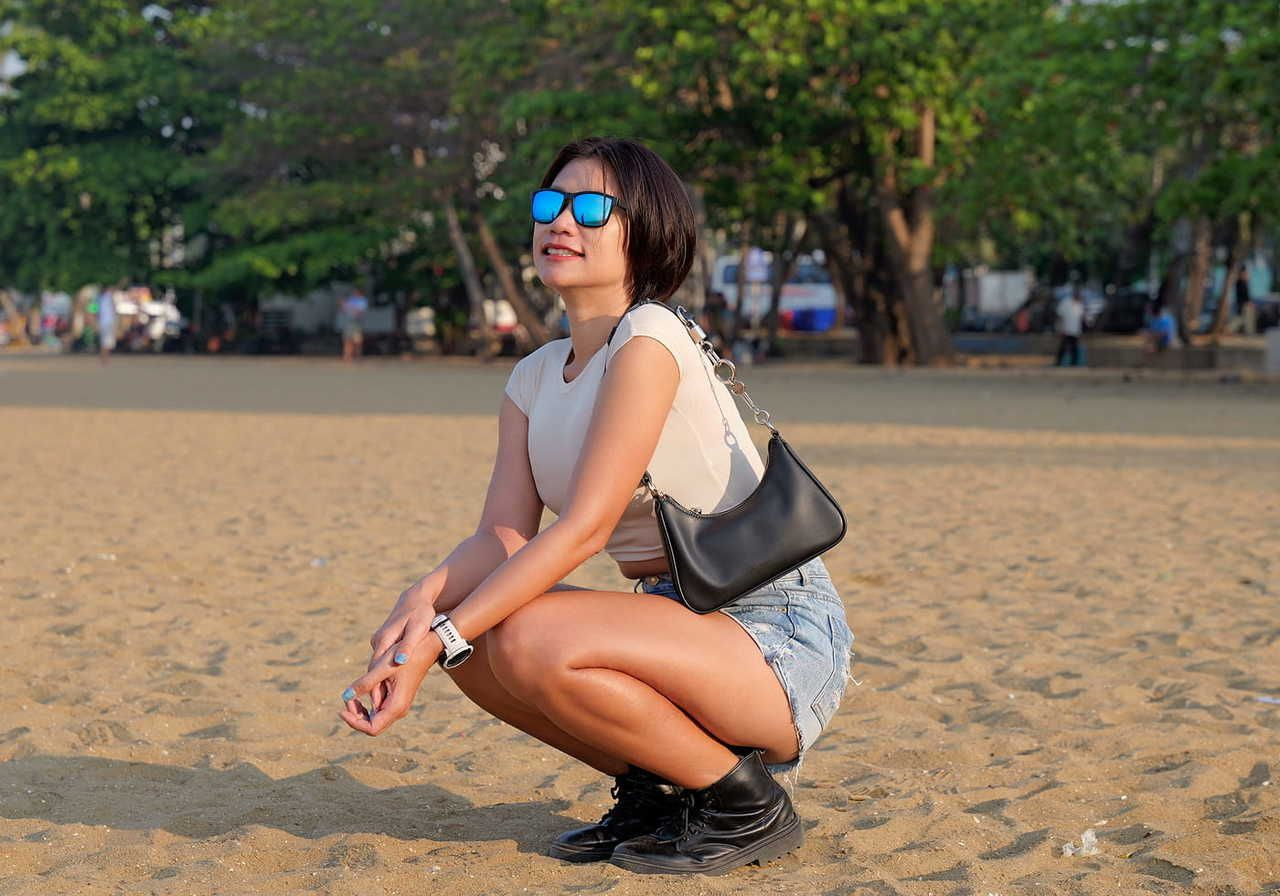 A woman squatting on the sand at a beach, wearing sunglasses, a white top, denim shorts, and black boots, with a black handbag.