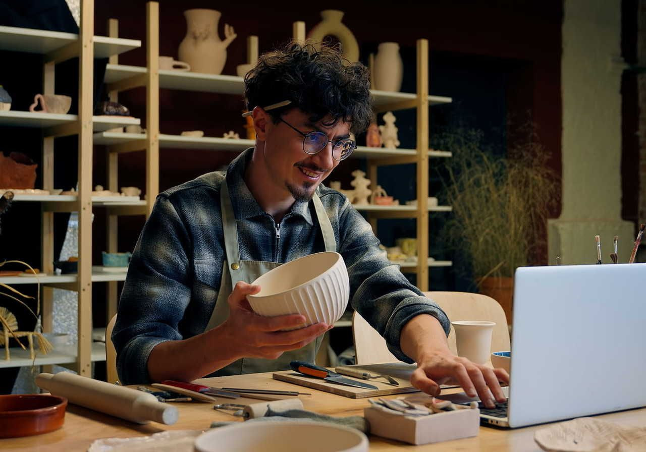 A smiling man sits at a laptop in a pottery studio, holding a ceramic bowl.