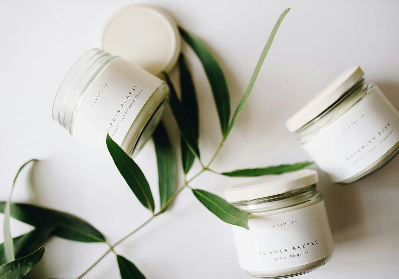 Three jars filled with white candles, surrounded by green leaves, arranged aesthetically on a neutral background.