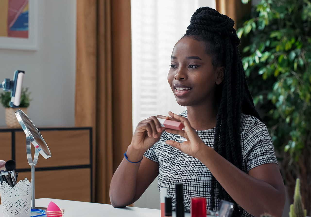 A woman with long braided hair holds a makeup item while filming a tutorial. She sits at a table with cosmetics in a bright, cozy room.