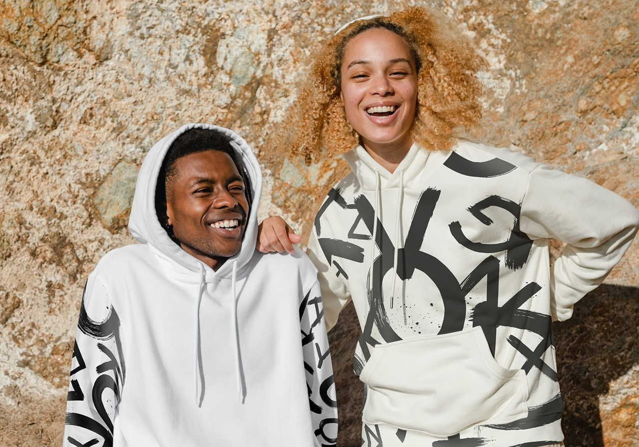 Two young individuals wearing hoodies with playful designs, standing against a rocky background and smiling at the camera.