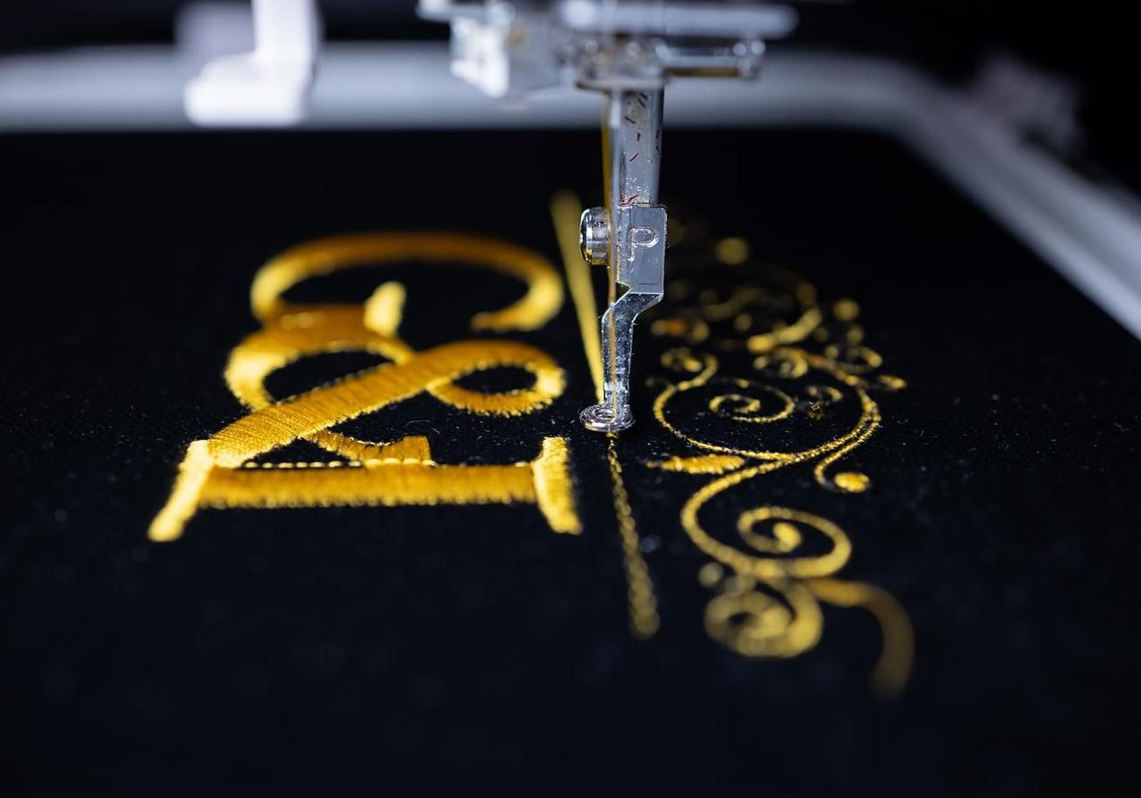 A sewing machine needle embroidering intricate gold patterns and letters on black fabric.