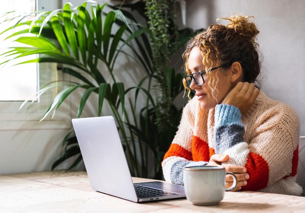 A woman sits at a table, wearing glasses and a colorful sweater, searching for the latest trends for Etsy sellers from a laptop.