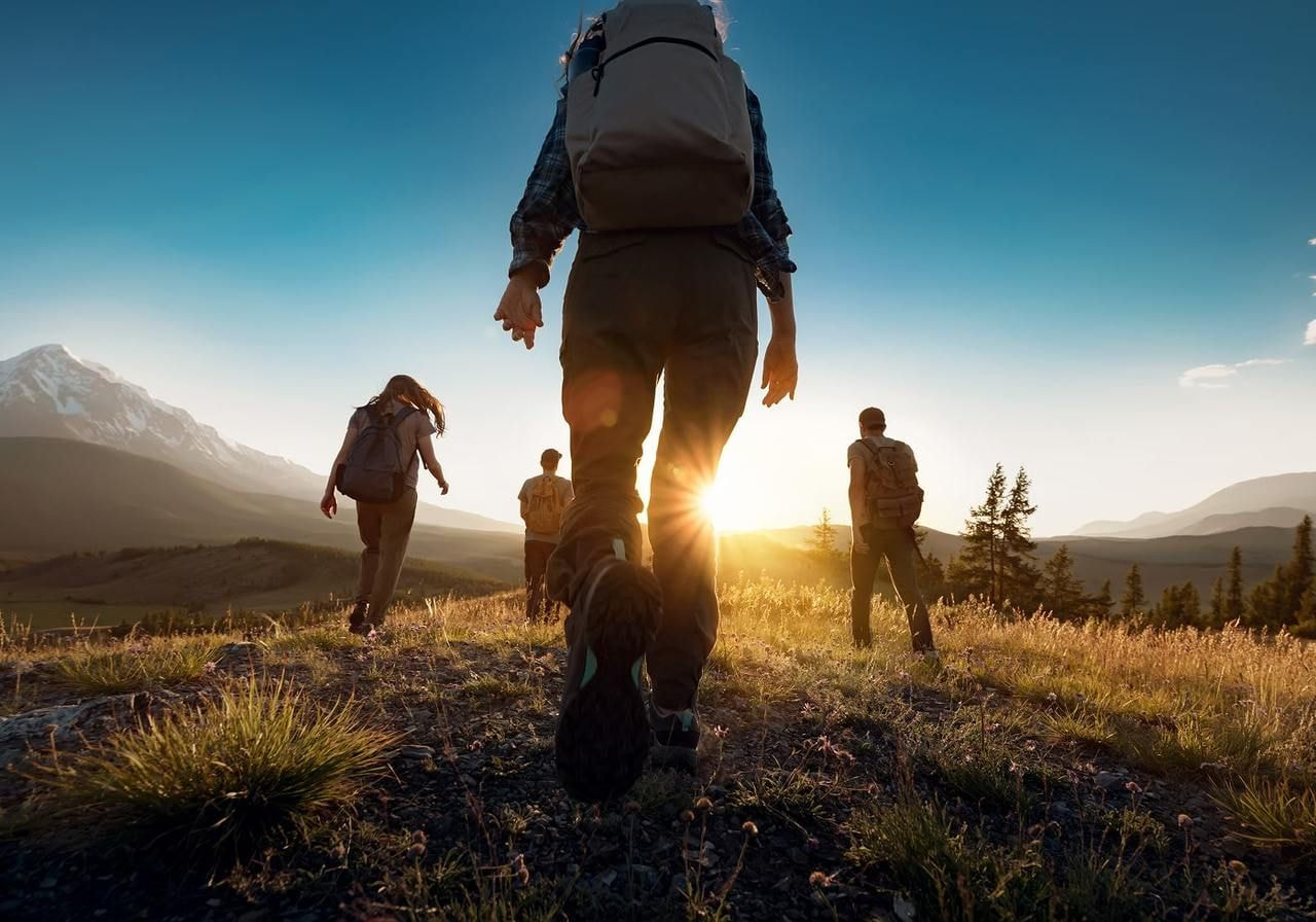 Hikers with backpacks walk through a sunlit mountain at sunrise, surrounded by distant peaks and trees.