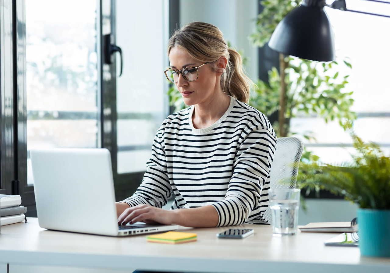 A woman with glasses, wearing a striped shirt, types on a laptop at a bright, plant-filled office desk.