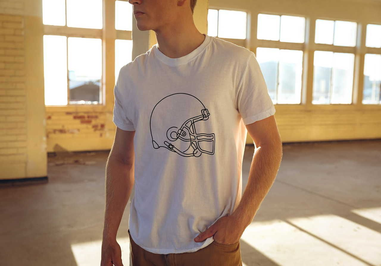 A man wearing a white t-shirt with a black outline of a football helmet stands in a sunlit room.