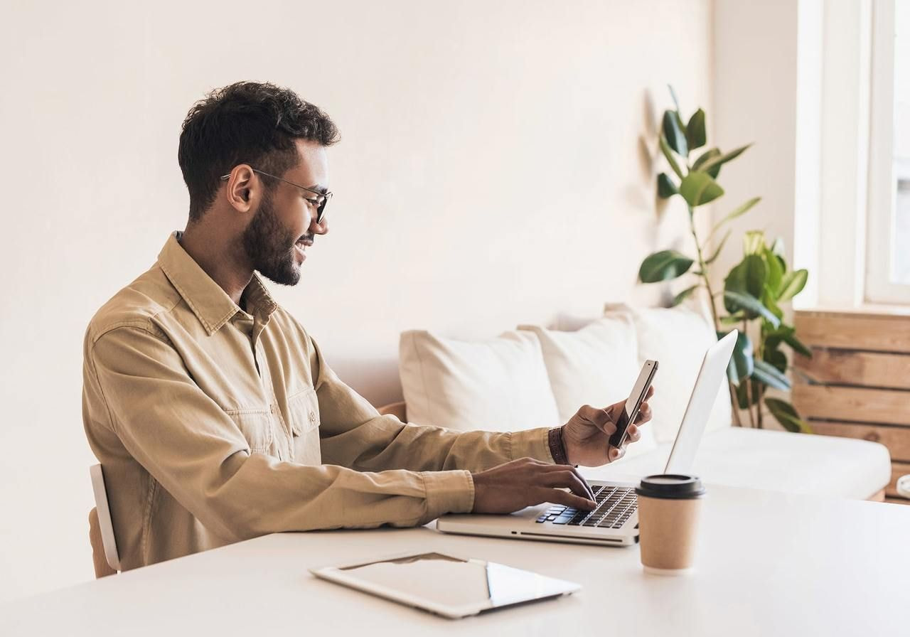 A man in a beige shirt is smiling while using a laptop and holding a smartphone, learning how much money can custom t-shirts make.