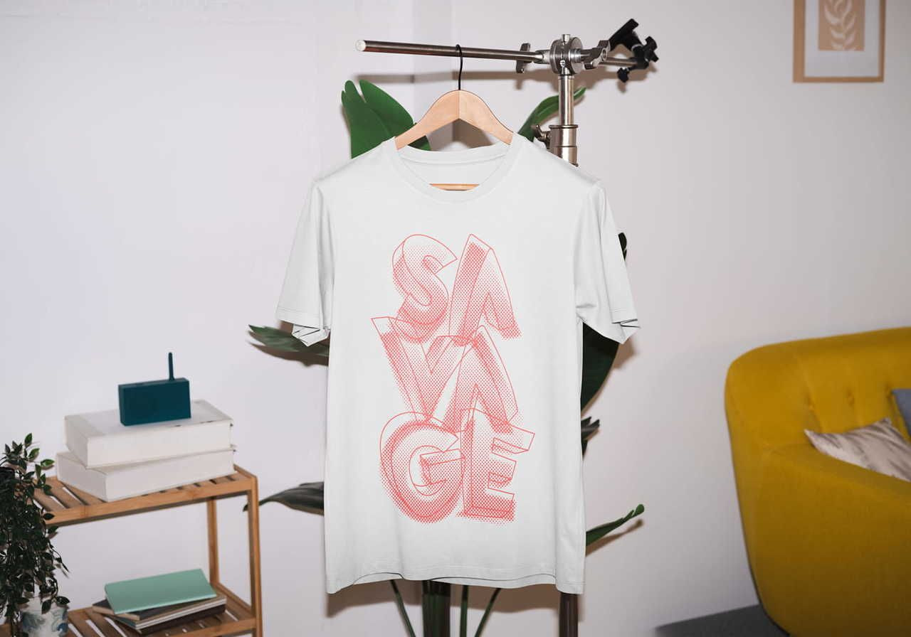 A white t-shirt with "SAVAGE" in bold red letters hangs on a stand. It's in a cozy room with plants, a yellow sofa, and a wooden shelf.