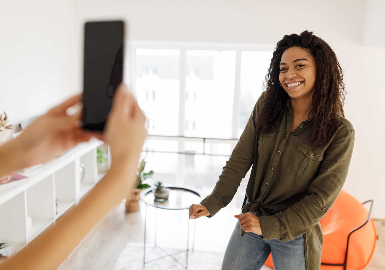 A smiling woman in a cozy living room dances playfully while being filmed with a mobile phone.