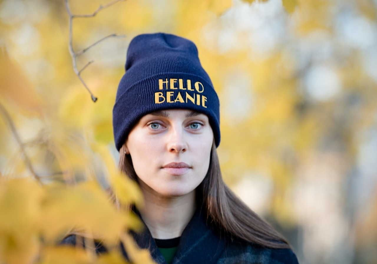 A young woman wearing a blue beanie with the text "HELLO BEANIE" against a backdrop of yellow leaves.
