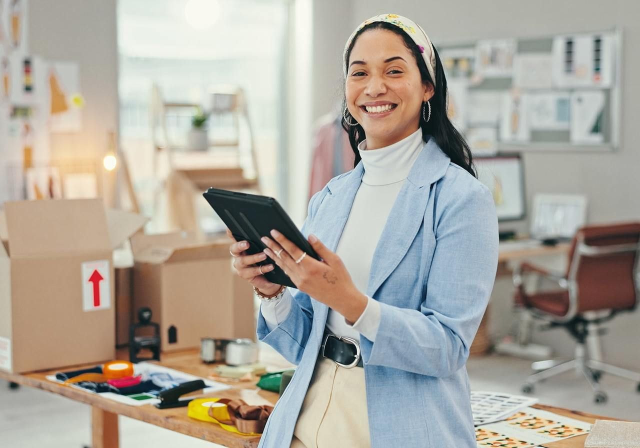 A smiling woman in a light blue blazer holds a tablet in a workspace. Open boxes and colorful materials on a table.