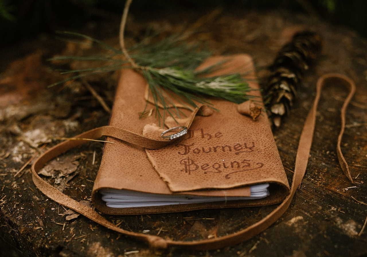 A rustic leather journal with "The Journey Begins" on the cover, adorned with pine needles, lies on a wooden surface next to a pinecone.