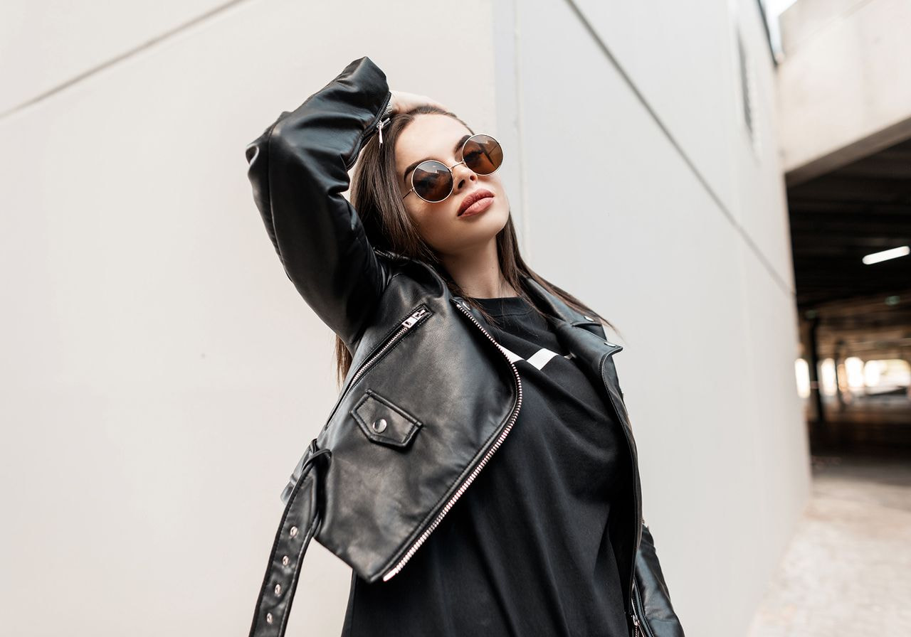 A stylish person poses confidently against a gray urban backdrop, wearing a black leather jacket and round sunglasses, exuding a cool, modern vibe.