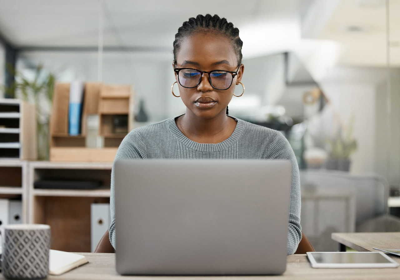 A woman in glasses and a grey sweater is sitting at a desk, focused on a laptop, learning how to start a print-on-demand business.