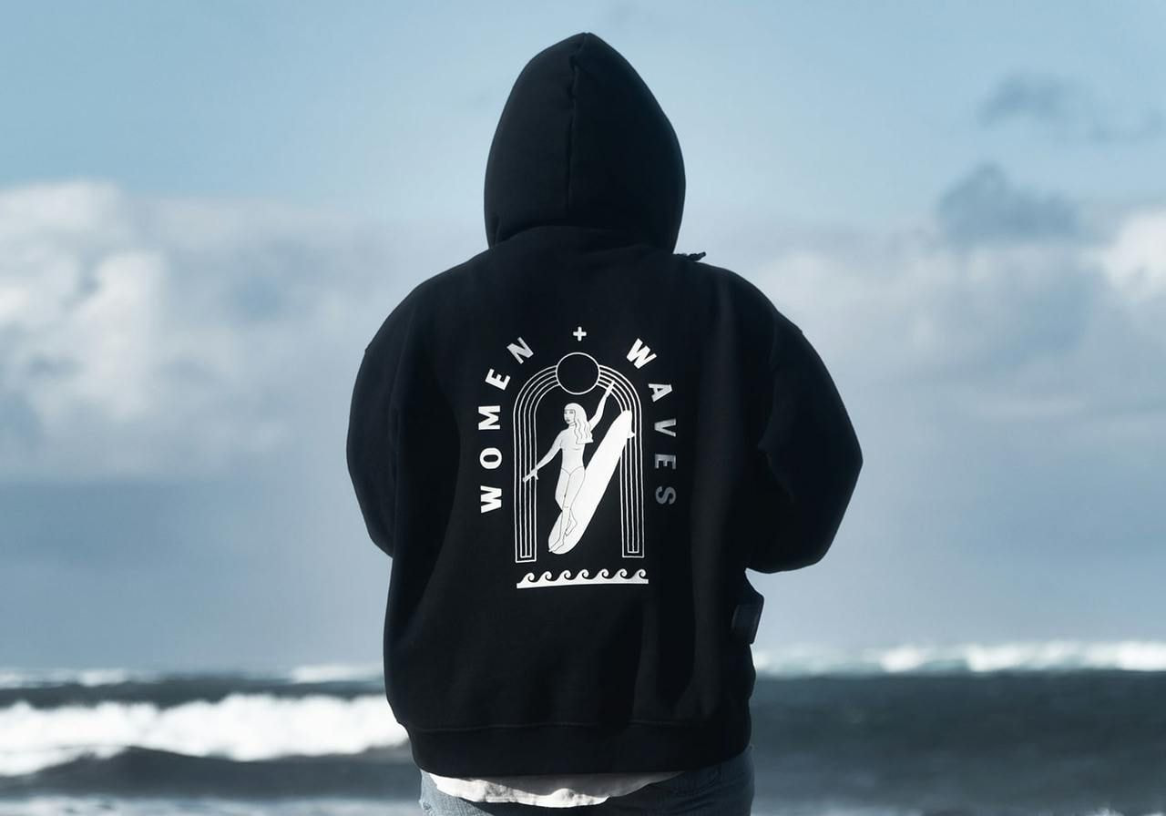 A backshot of a woman in a blue “Women + waves” hoodie standing in front of the sea.