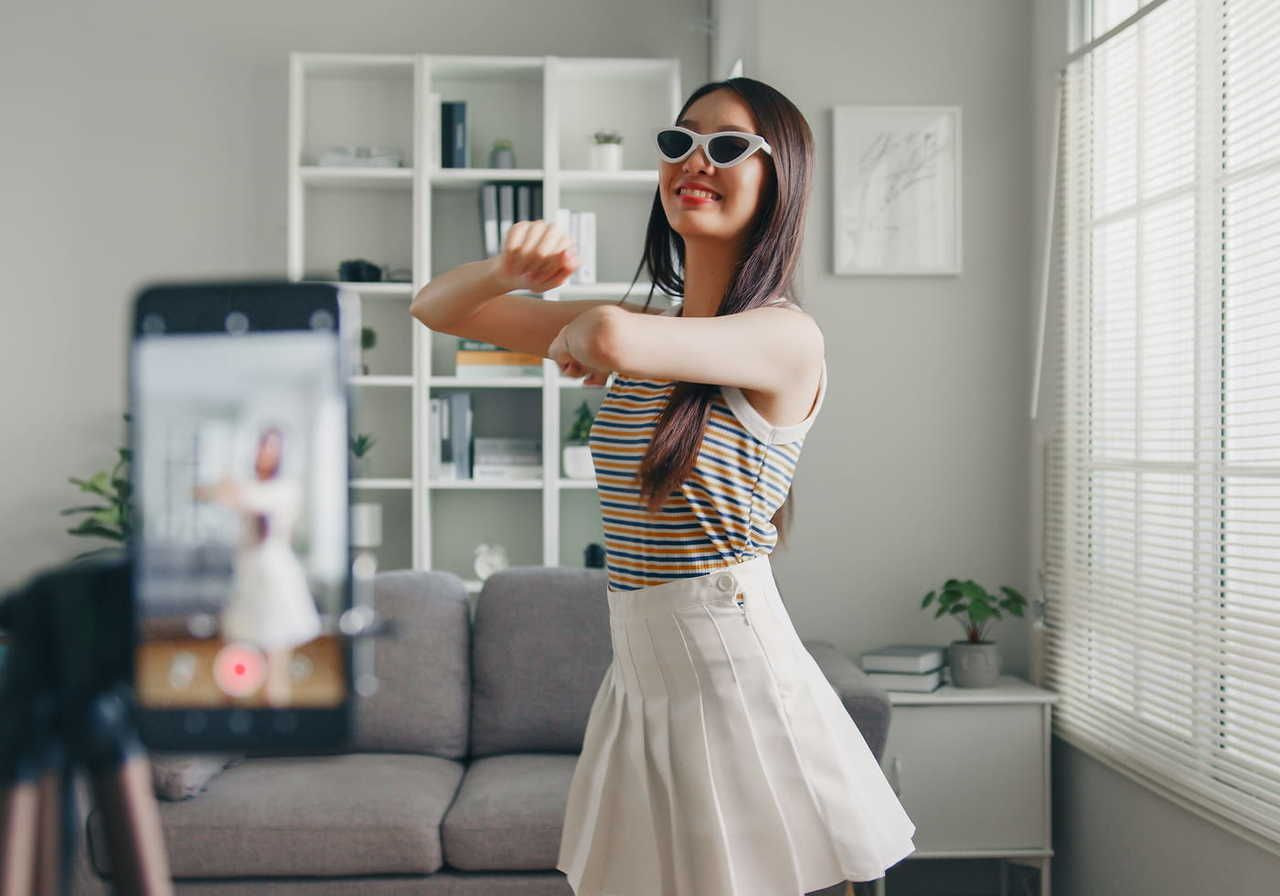 A young woman in sunglasses dances while recording herself on a smartphone in a modern living room.