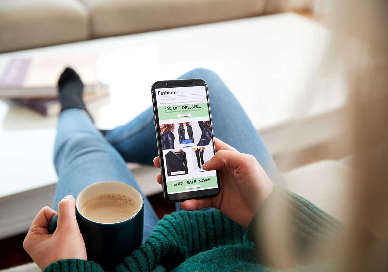 A man relaxes on a couch, browsing online fashion deals on a smartphone. He holds a mug, wearing jeans and a green sweater.